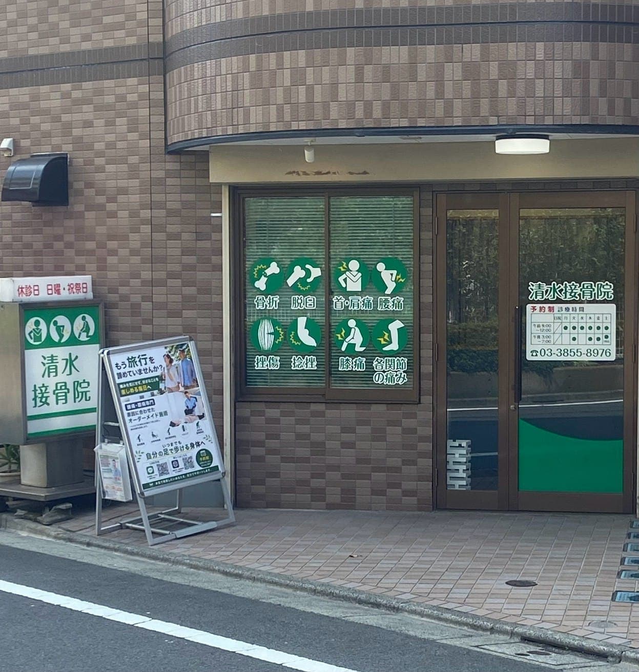 清水接骨院の外観。緑の看板と、腰痛・膝痛など対応症状のイラストが描かれた窓が目印